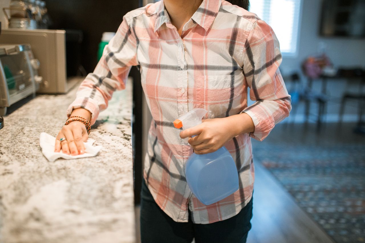 hero-img A woman wearing a plaid shirt cleans a kitchen counter using a spray bottle and cloth.